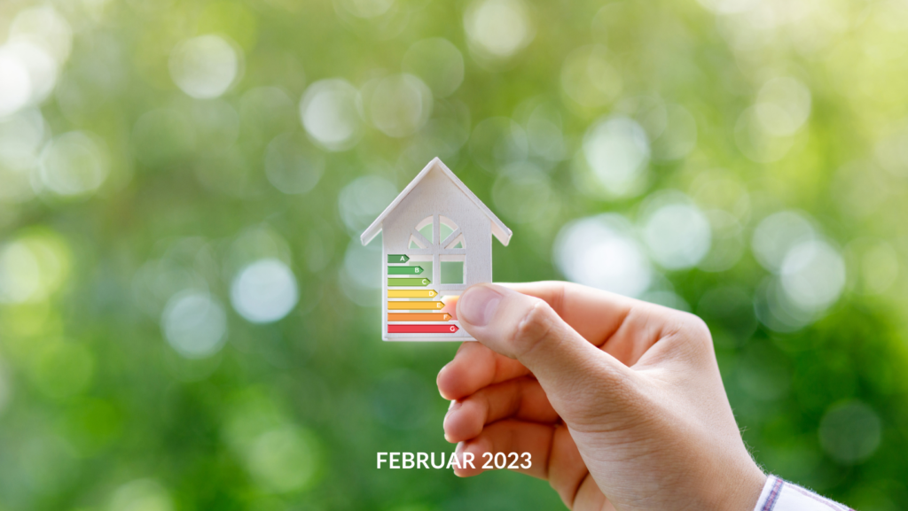 Eine Hand hält in der Immobilienbranche vor einem unscharfen grünen Hintergrund ein kleines hausförmiges Symbol mit einer bunten Energieeffizienzskala und der Aufschrift „Februar 2023“.