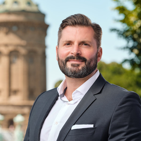 Porträt eines lächelnden bärtigen Mannes in dunklem Anzug und weißem Hemd, der im Freien steht, im Hintergrund Britta Schentke-Becker.