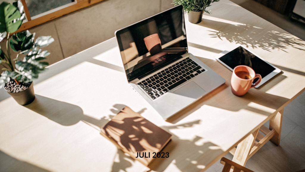 Ein sonnendurchfluteter hybrider Arbeitsplatz mit einem aufgeklappten Laptop, einem Notizbuch, einer Tasse und Pflanzen auf einem Holztisch mit der Aufschrift „Juli 2023“.