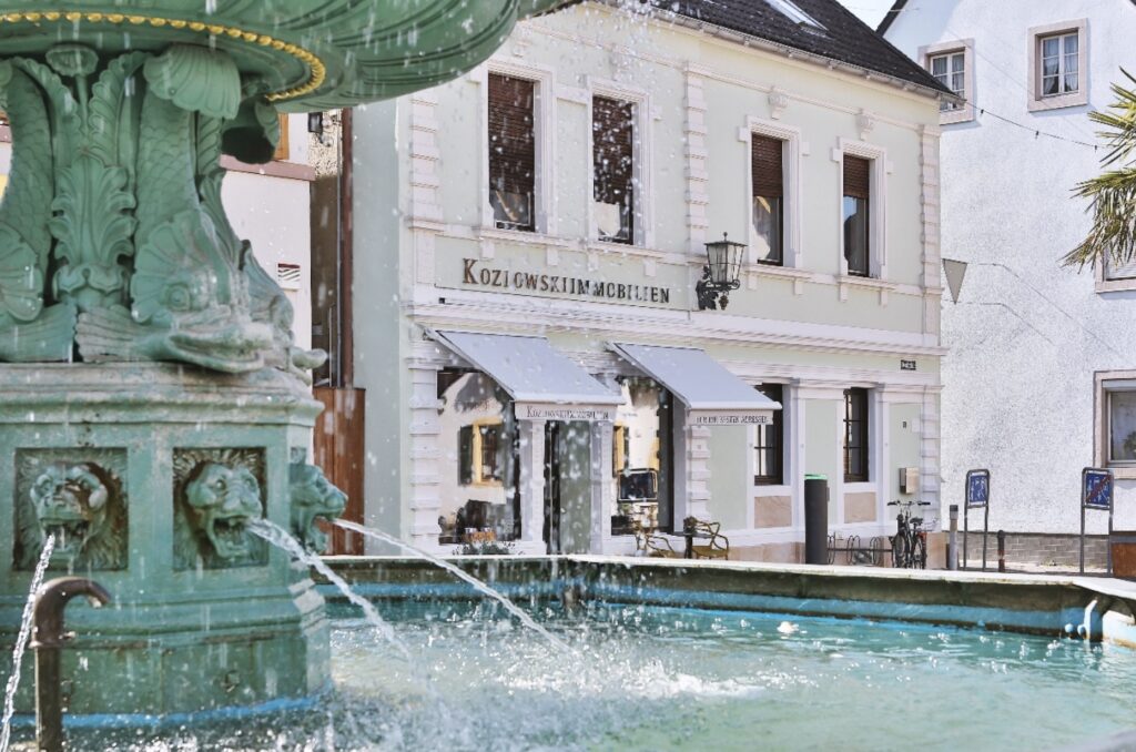 Ein Brunnen mit Wasser spritzenden Löwenkopf-Skulpturen auf einem malerischen Stadtplatz, im Hintergrund ein Gebäude mit der Aufschrift „kozlovski smčdlien“.