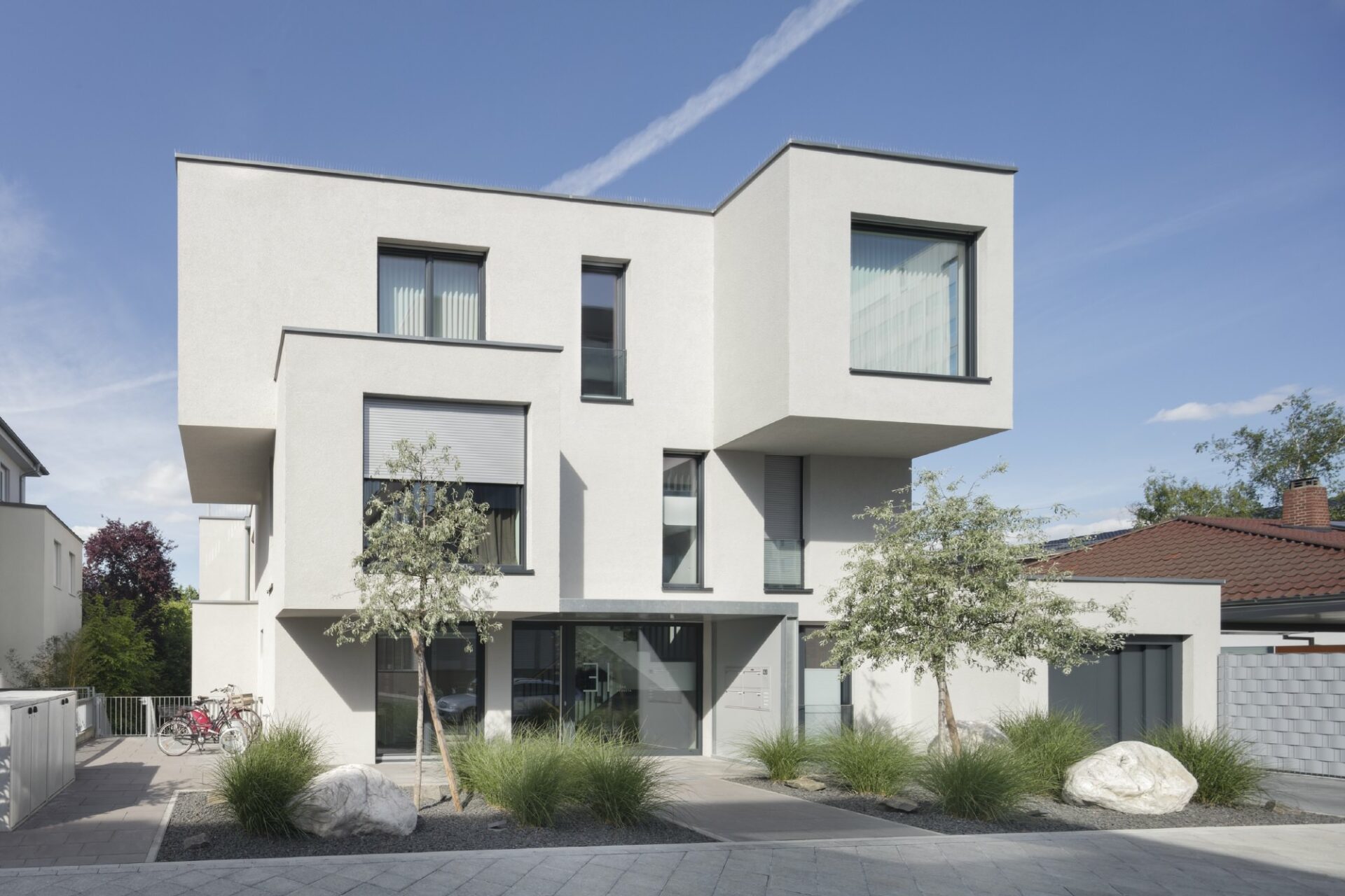 Modernes weißes Haus in kubischer Architektur mit großen Fenstern, umgeben von einem angelegten Garten und klarem blauen Himmel.