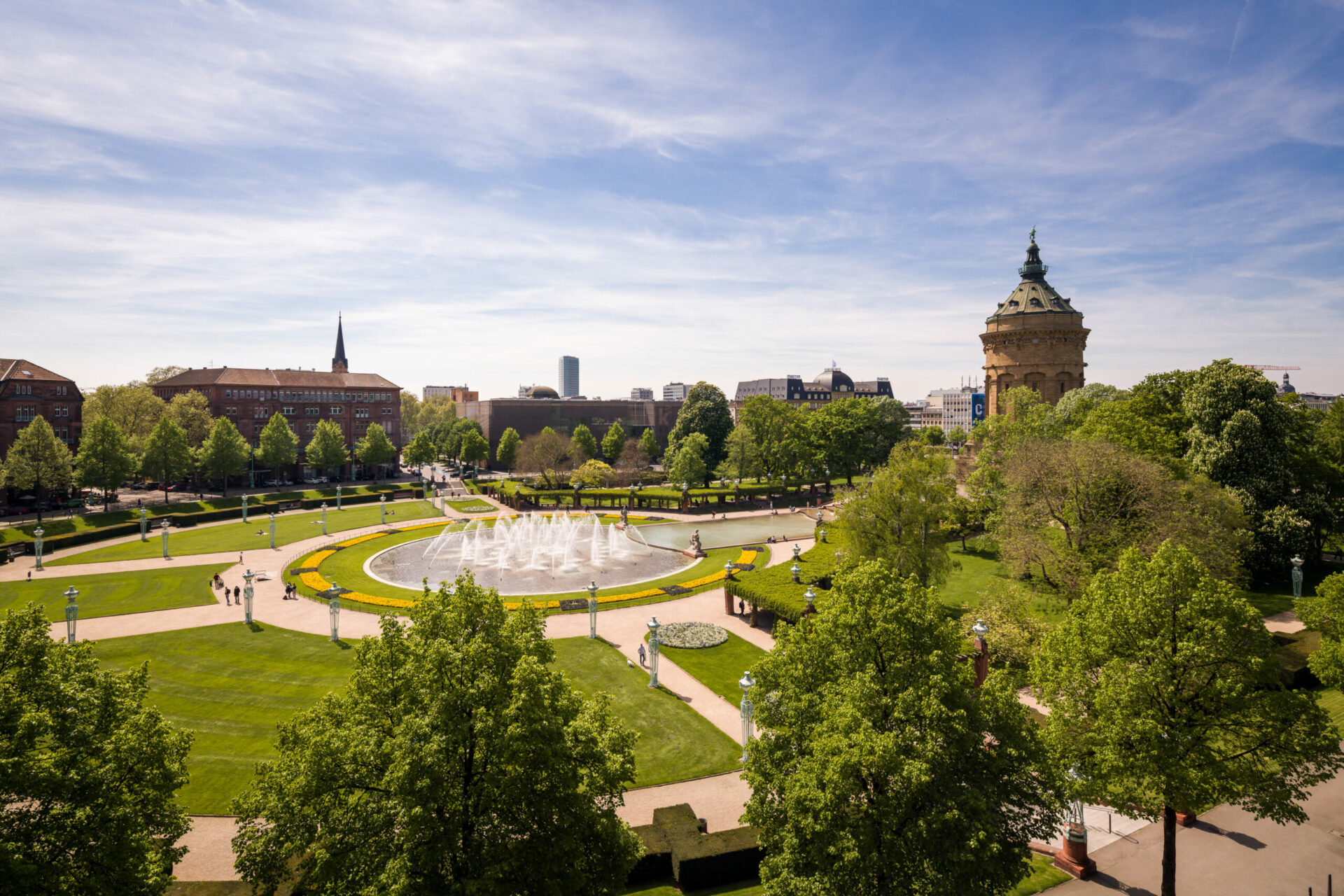 Luftaufnahme des Rheinauparks mit einem großen Brunnen in der Mitte, umgeben von gepflegten Rasenflächen und Wegen, mit historischen Gebäuden unter einem klaren blauen Himmel.