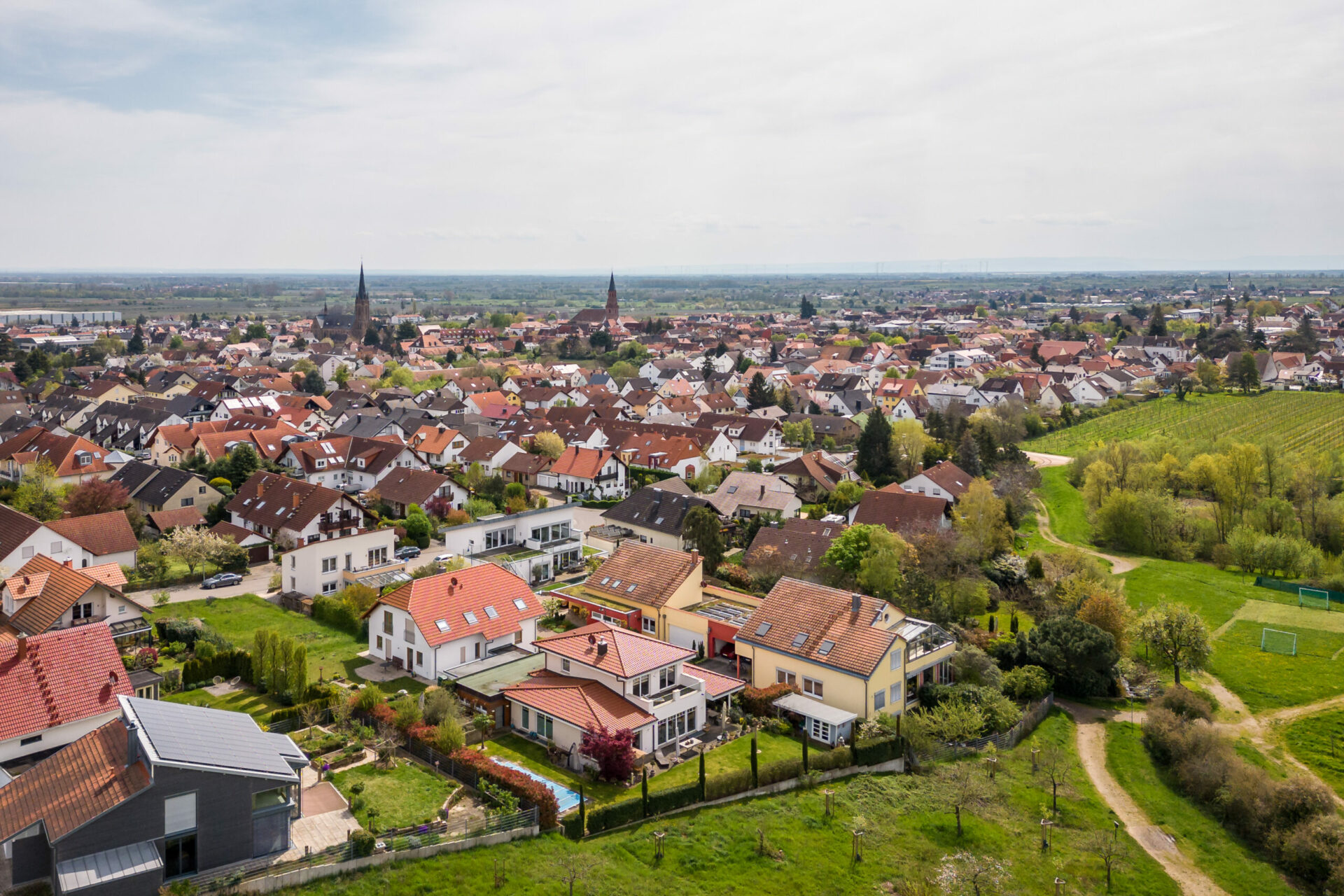 Luftaufnahme von Edenkoben mit angesiedelten Häusern, Straßen und Grünflächen, im Hintergrund ist die Stadt unter einem bewölkten Himmel zu erkennen.