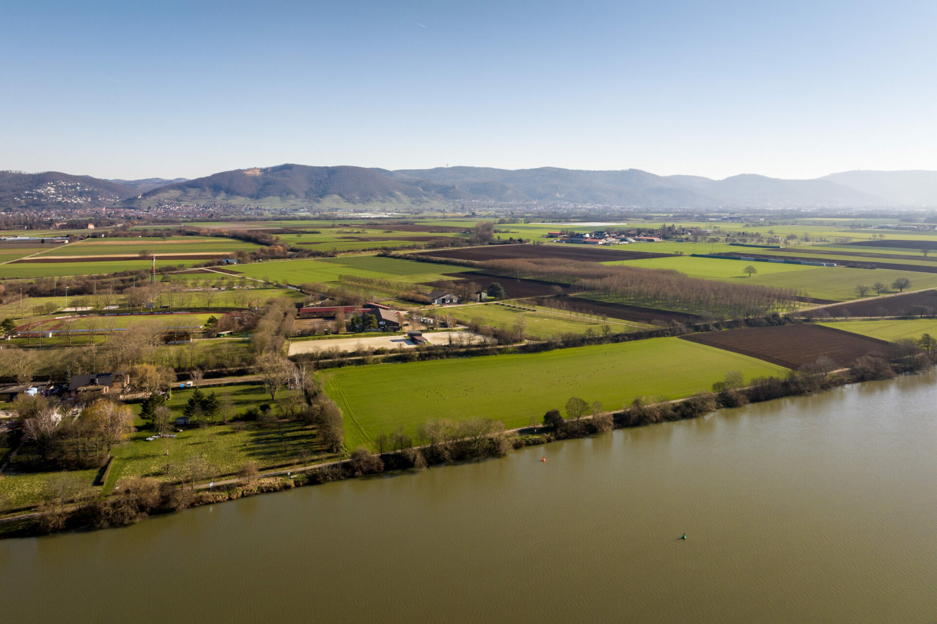 Luftaufnahme eines ruhigen Flusses neben üppigen grünen Feldern und verstreuten Gebäuden in Edingen-Neckarhausen, mit Hügeln in der Ferne unter einem klaren blauen Himmel.