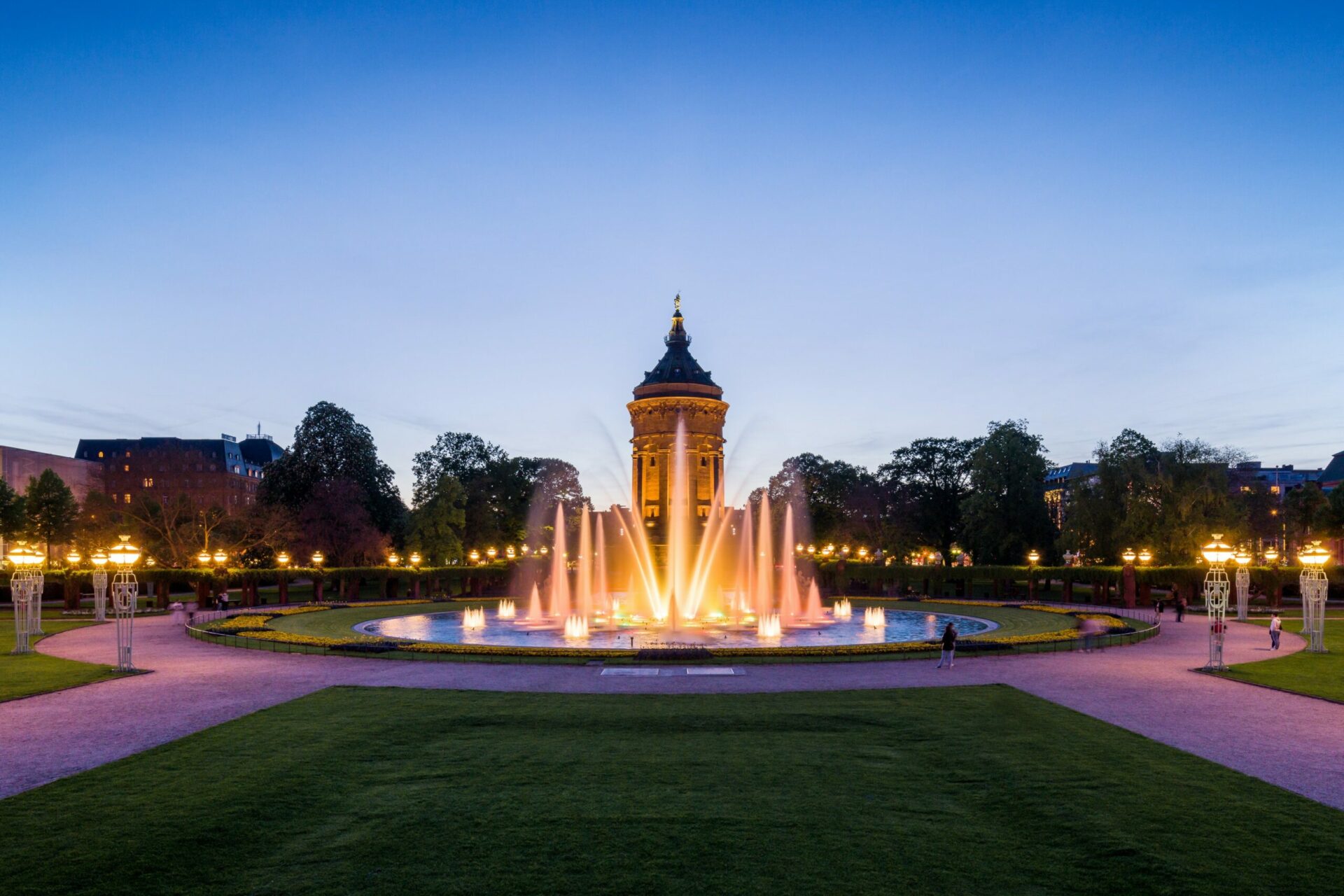 Ein beleuchteter Brunnen und ein Pavillon in der Abenddämmerung im Birkweiler Park, mit blauem Himmel und Stadtgebäuden im Hintergrund.