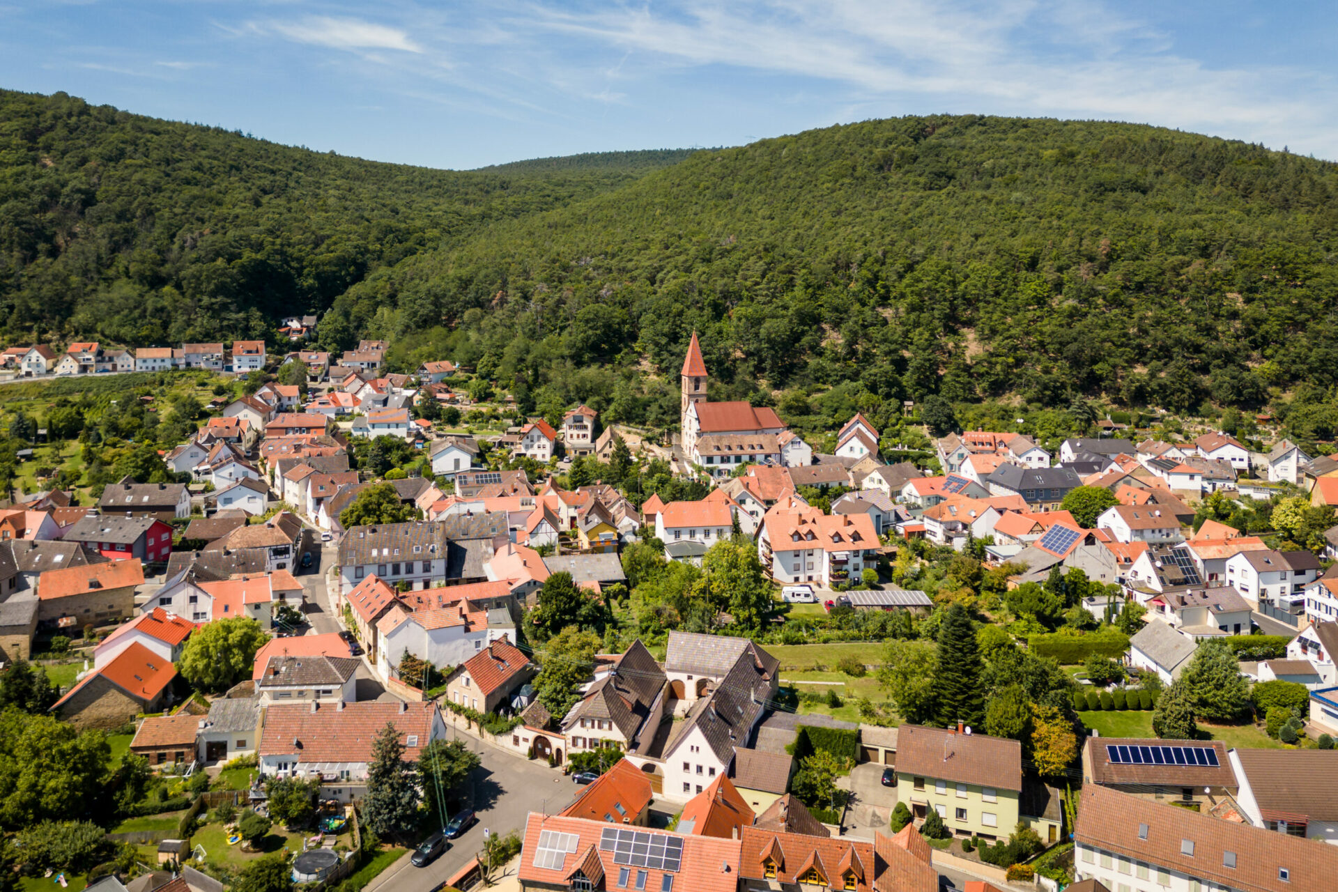 Luftaufnahme des malerischen Dorfes Königsbach mit dicht gedrängten Häusern, einem markanten Kirchturm und umgeben von üppigen bewaldeten Hügeln.