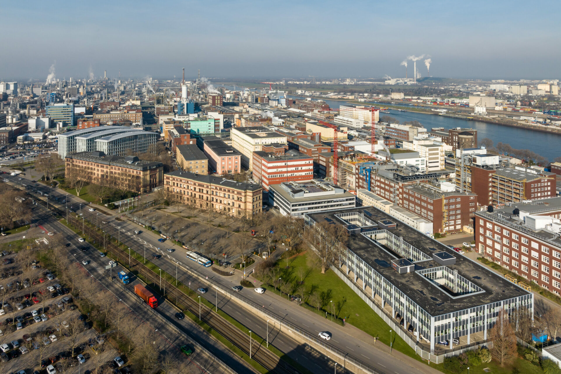 Luftaufnahme einer modernen Stadtlandschaft in Ludwigshafen, Deutschland, mit Gebäudegruppen entlang eines Flusses, Straßen und klarem blauen Himmel.