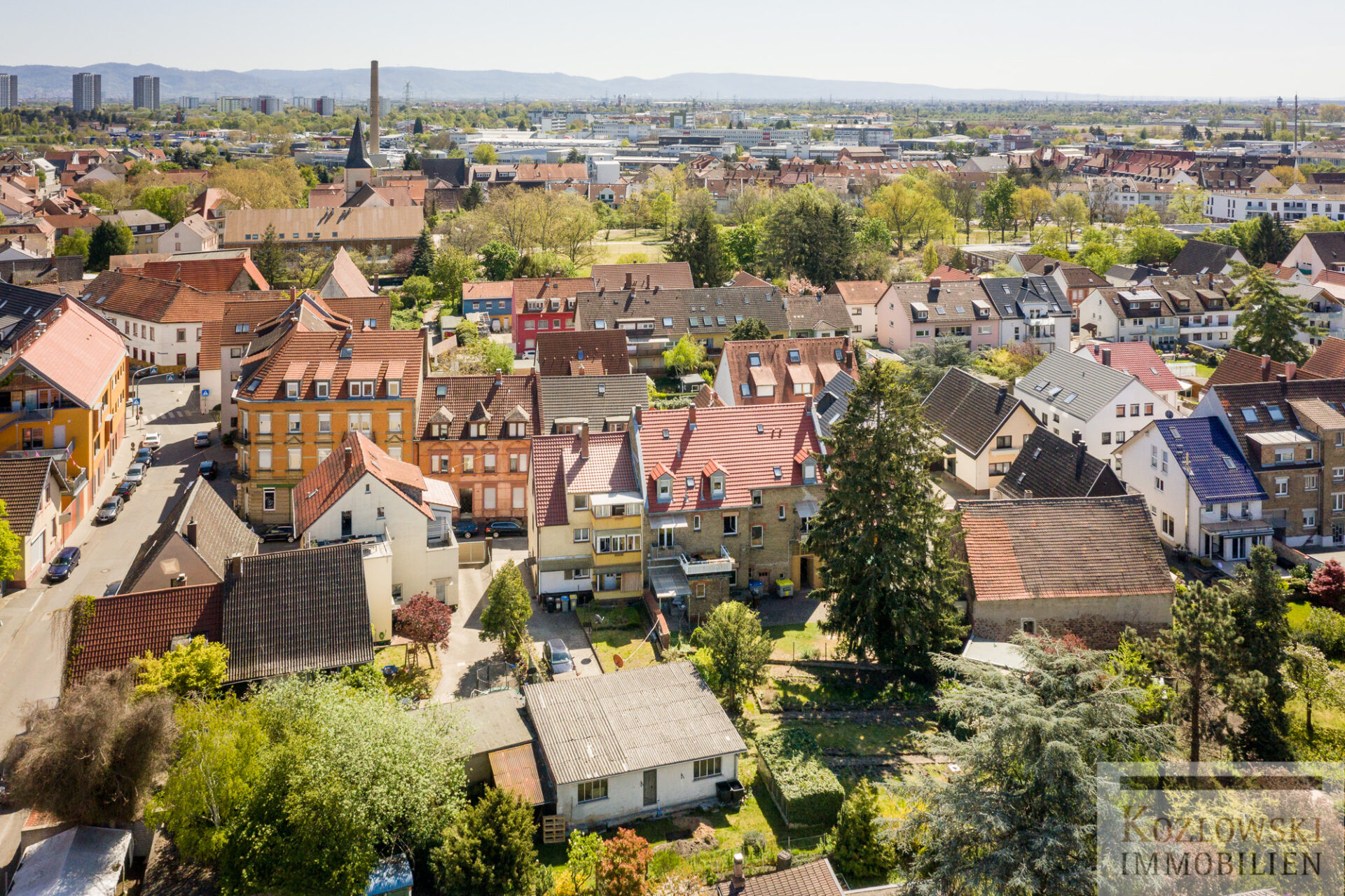 Luftaufnahme des Vorortgebiets Käfertal. Zu sehen sind Reihen von Häusern mit markanten roten und grauen Dächern, durchzogen von Straßen, mit einer Stadtlandschaft im Hintergrund.