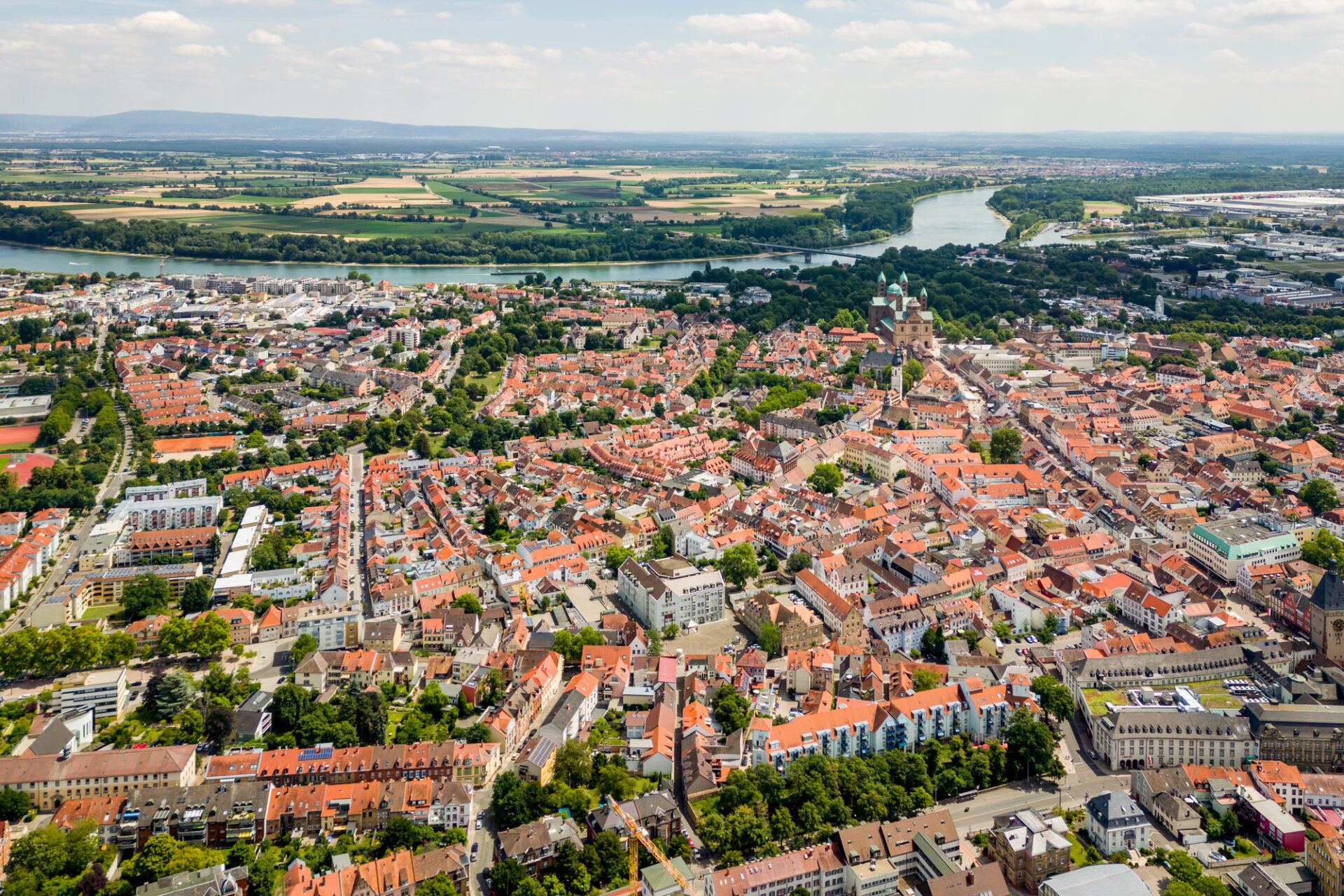 Luftaufnahme einer dicht besiedelten deutschen Stadt mit roten Dächern, durch die der Rhein fließt und die von grünen Feldern umgeben ist.