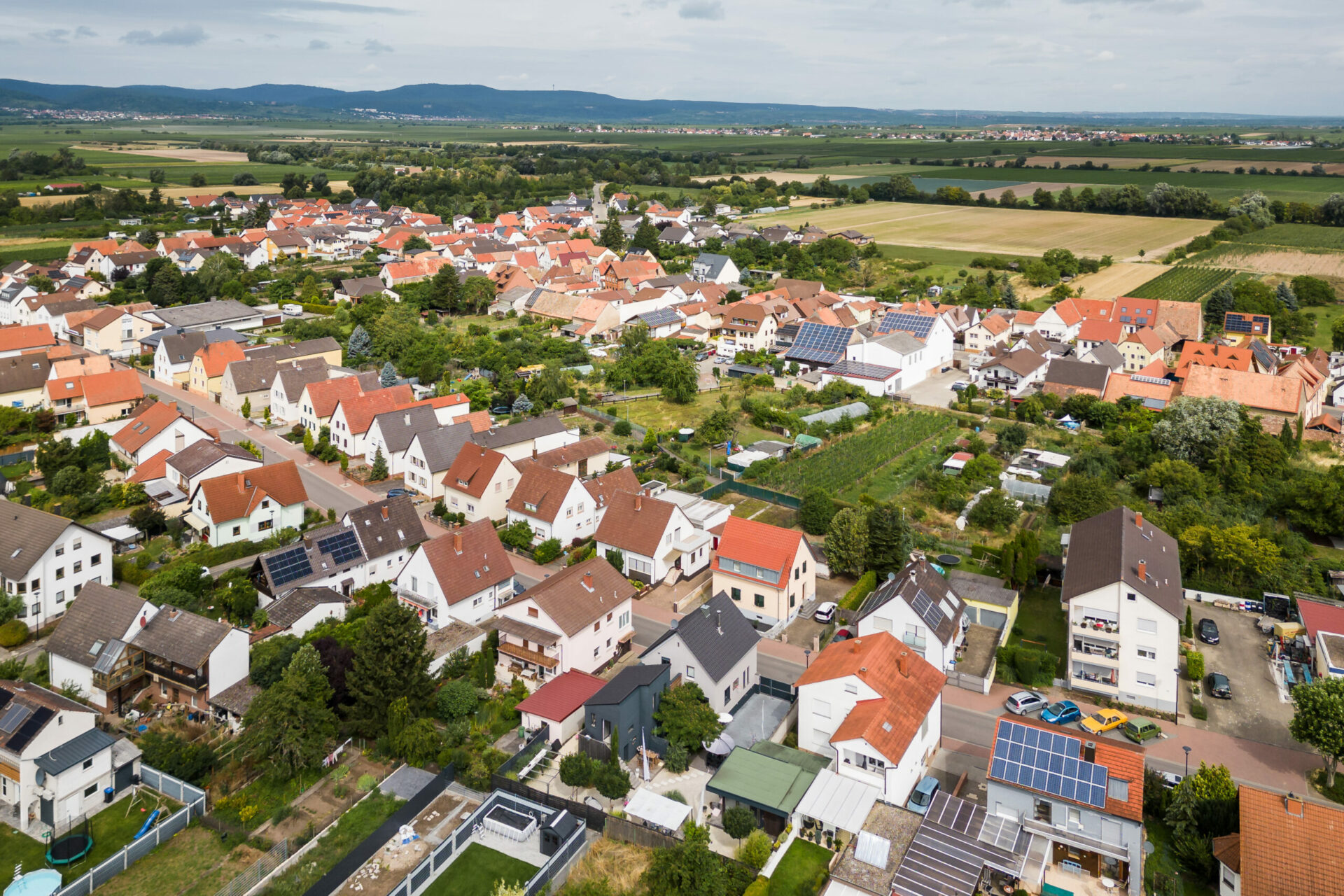 Luftaufnahme des Stadtteils Rödersheim-Gronau mit dicht gedrängten Häusern, teilweise mit Solarmodulen, umgeben von Feldern und in der Ferne Hügeln.