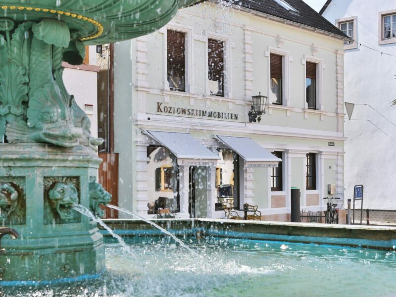 Ein lebhafter Stadtplatz mit einem detailreichen grünen Brunnen im Vordergrund und charmanten Gebäuden im europäischen Stil im Hintergrund.