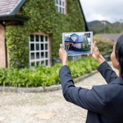 Eine Frau im Businessanzug hält Vermietung das Foto eines Hauses hoch und richtet es am tatsächlichen Haus im Hintergrund aus.