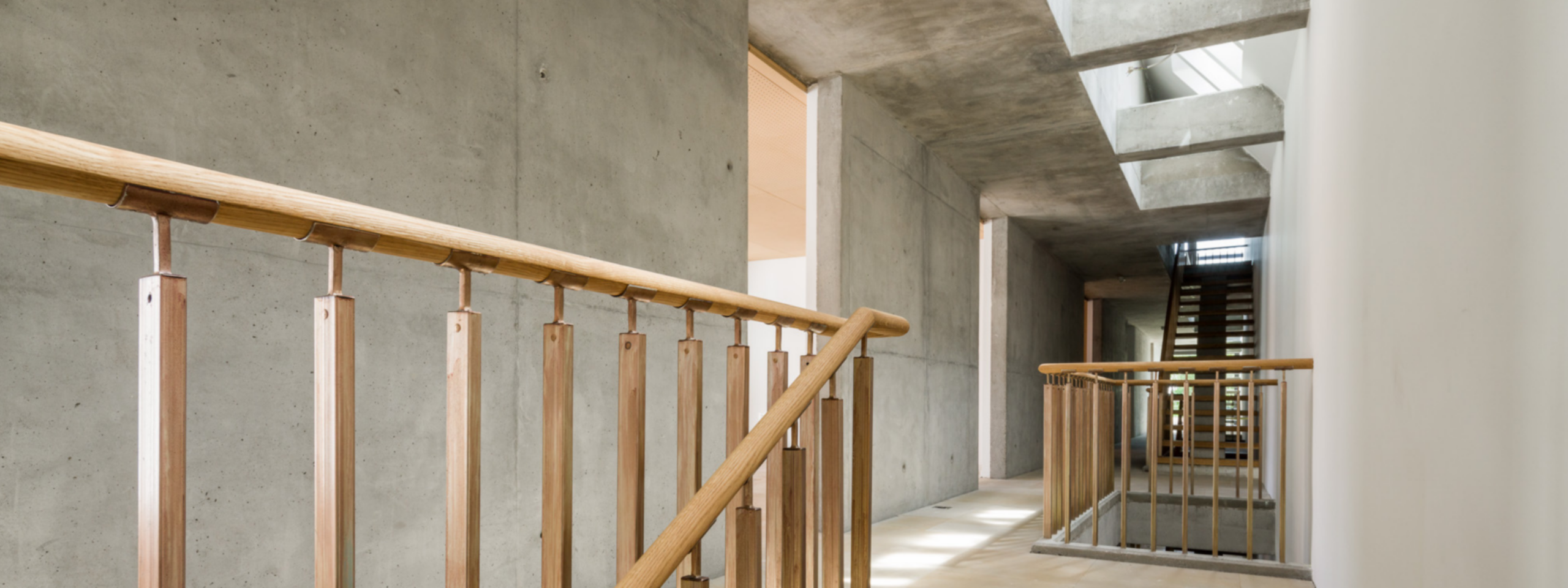 Innenansicht eines modernen Gebäudes mit freiliegenden Betonwänden und Holzhandläufen entlang einer Treppe und eines Balkons.