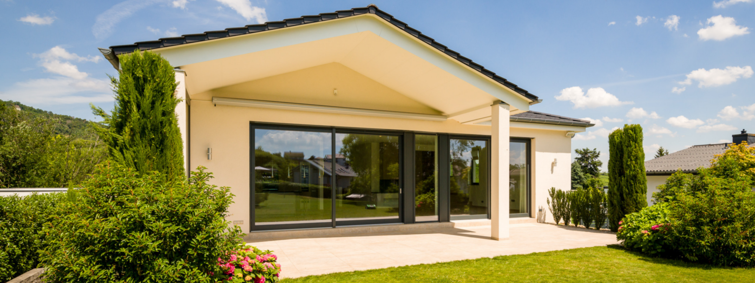 Modernes einstöckiges Haus mit großen Glasfenstern und beigen Wänden, umgeben von einem gepflegten Rasen und leuchtenden Blumenbüschen unter einem klaren Himmel.