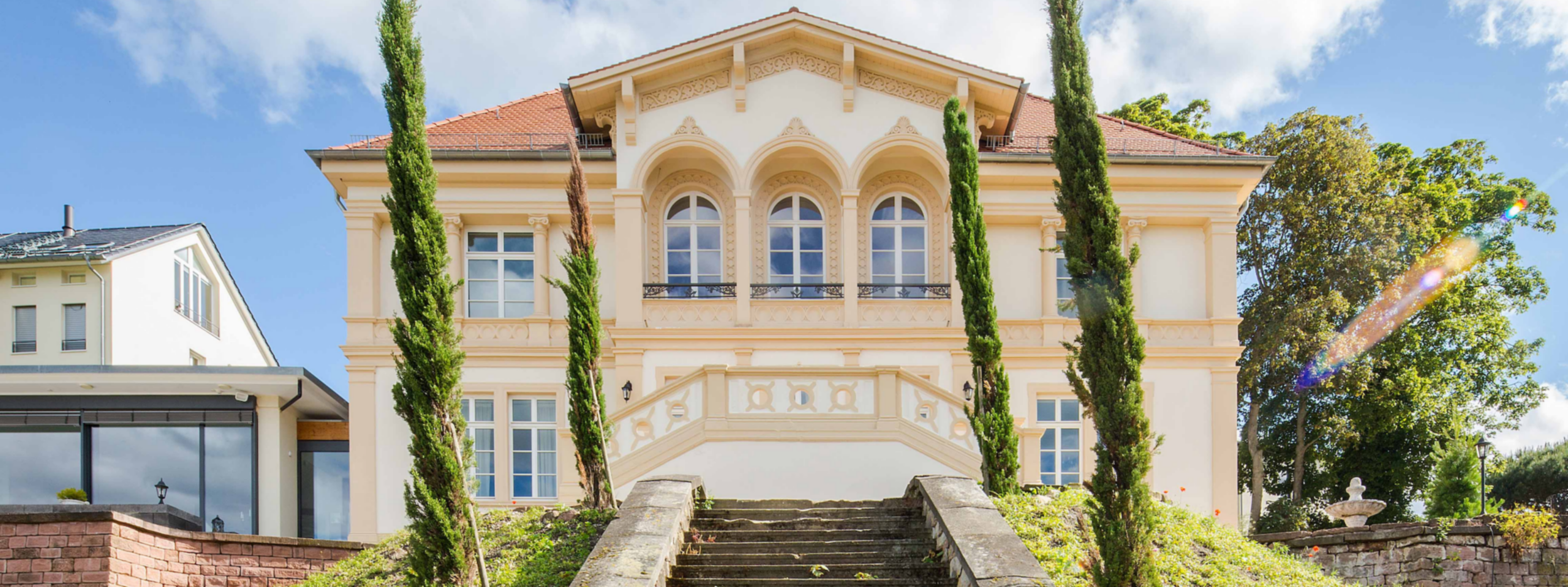 Aufwändig gestaltetes, hellgelbes Herrenhaus mit großem Eingang, flankiert von zwei Zypressen unter einem sonnigen Himmel.
