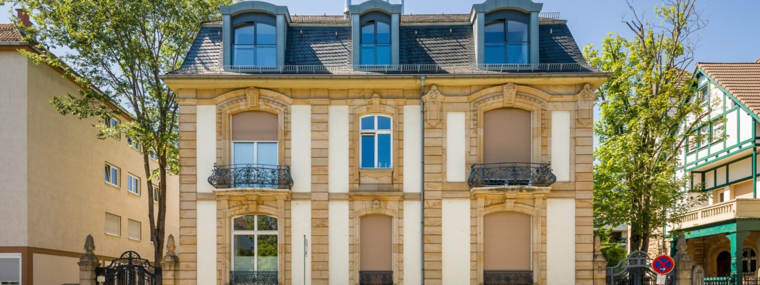 Elegantes zweistöckiges Gebäude mit symmetrischem Design, Bogenfenstern, Balkonen und Mansardendach, in einer sonnigen Stadtstraße.
