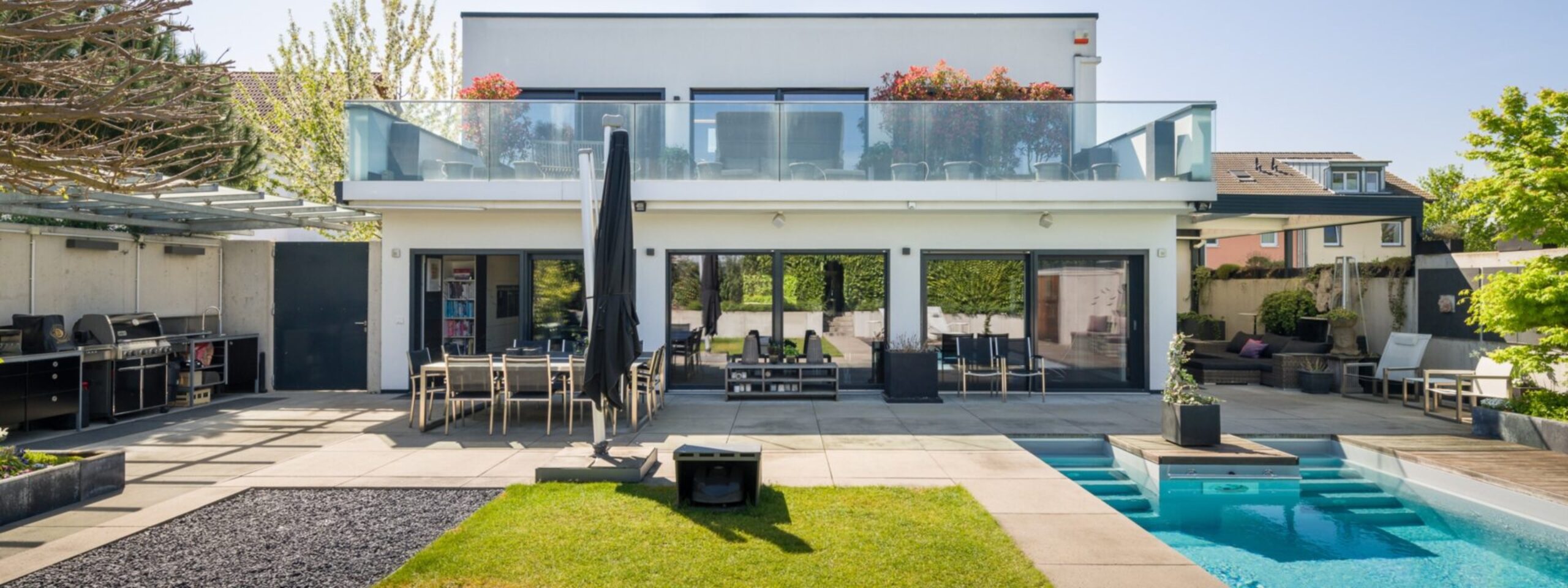 Modernes zweistöckiges Haus mit großen Fenstern, einem Balkon und einem Außenbereich mit Swimmingpool, Gartenmöbeln und angelegtem Garten.