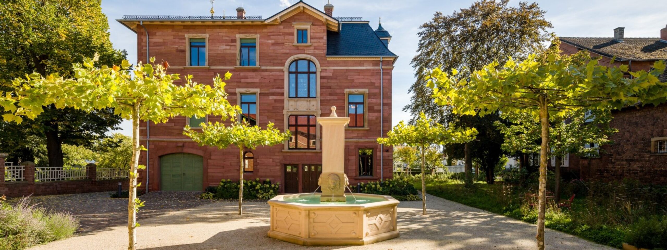 Ein gepflegtes Sandsteinhaus mit grüner Tür, flankiert von üppigen Bäumen und einem kleinen Brunnen im Vordergrund.