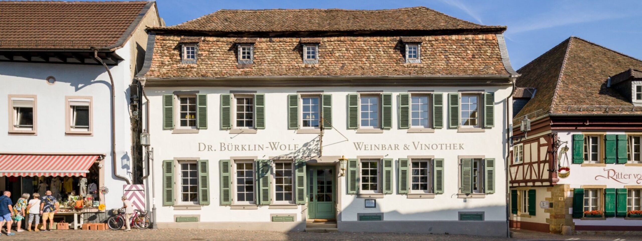 Historische europäische Straße mit traditionellen Gebäuden, darunter eines mit der Aufschrift „Dr. Bürklin-Wolf Weinbar & Vinothek“, unter einem klaren blauen Himmel.