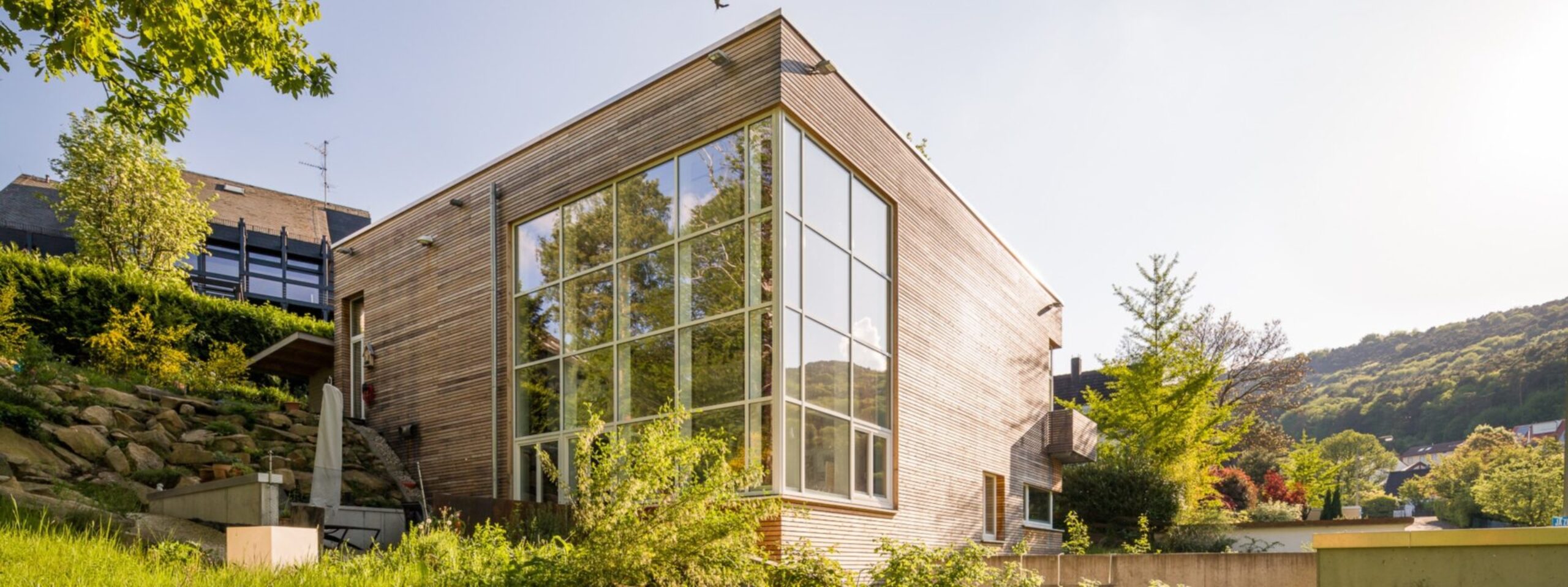 Modernes eckiges Haus mit großen Glasfenstern, umgeben von Grün unter einem klaren Himmel.