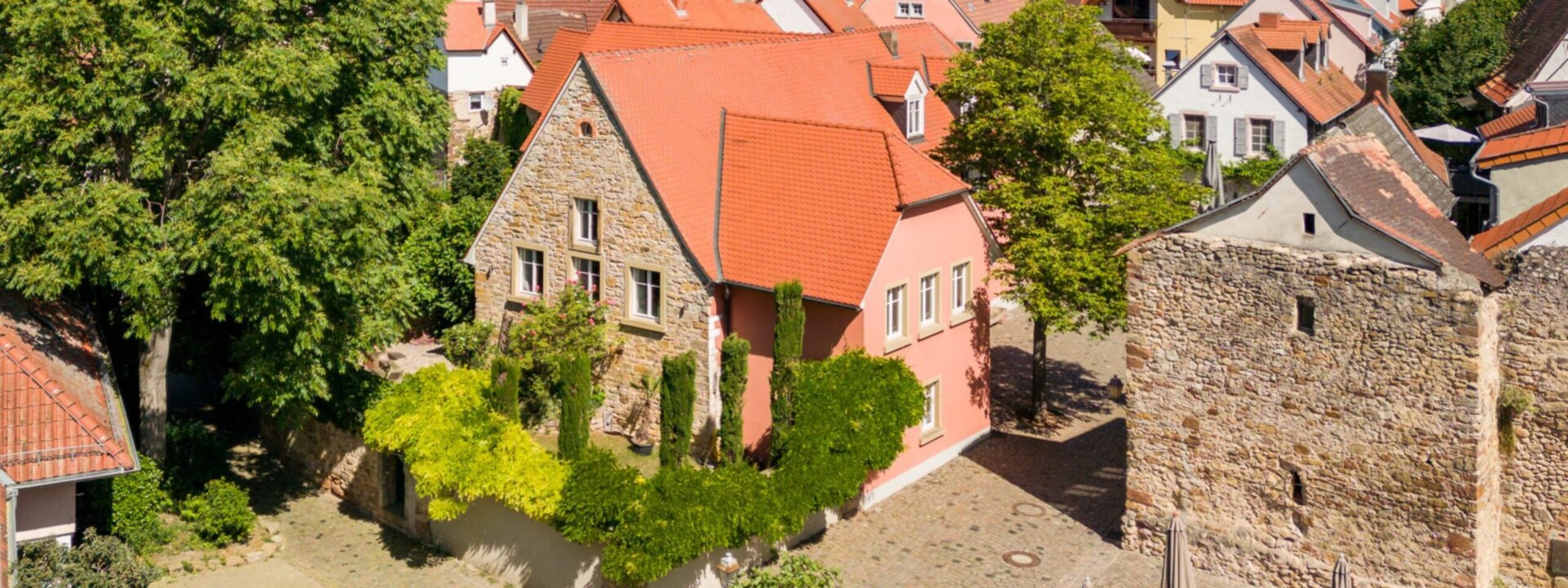 Luftaufnahme eines europäischen Dorfes mit traditionellen Häusern mit roten Ziegeldächern, umgeben von alten Steinmauern und üppigen Bäumen.