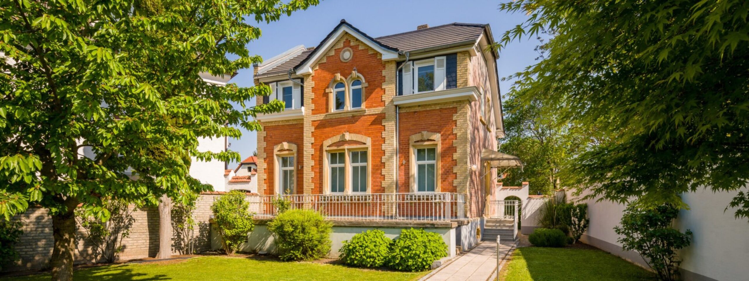 Ein zweistöckiges Backsteinhaus mit großen Fenstern und einem Balkon, umgeben von einer grünen Landschaft und einem klaren Himmel.