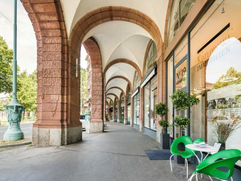 Gewölbter Gehweg mit roten Steinsäulen, grünen Stühlen in einem Café und Geschäften entlang einer Stadtstraße an einem sonnigen Tag.