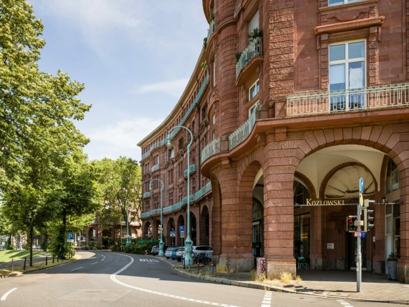 Rotes Backsteingebäude mit gewölbten Eingängen an einer Straßenecke, grünen Balkonen aus Schmiedeeisen und einem klaren blauen Himmel.